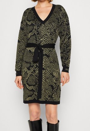 Mujer lleva un vestido de punto negro y dorado con patrón hasta la rodilla, con escote en V, mangas largas, cinturón negro en la cintura y botas negras hasta la rodilla.