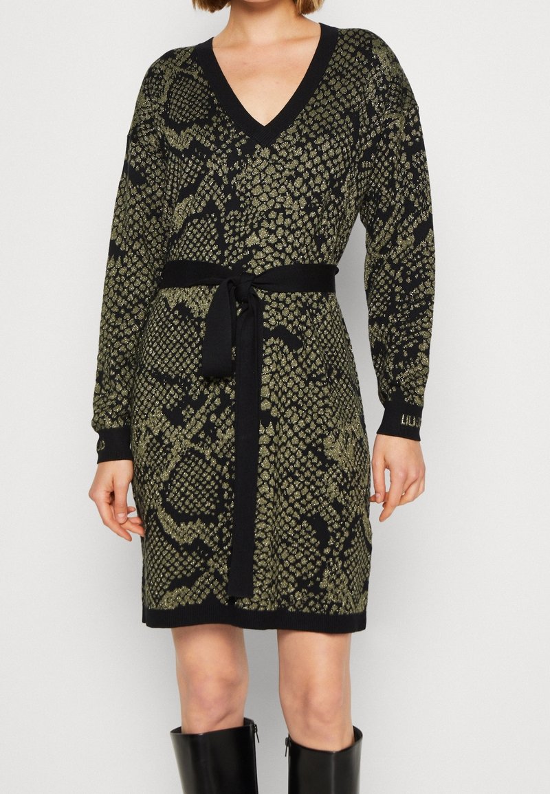 Une femme porte une robe en tricot noire et dorée à motifs, longueur genou, avec un col en V, des manches longues, une ceinture noire à la taille, et des bottes noires montant jusqu'aux genoux.