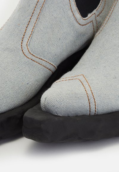 Zapatos de lona azul con detalles de costura naranja, punta redonda y suela gruesa de goma negra, con un acabado texturizado.