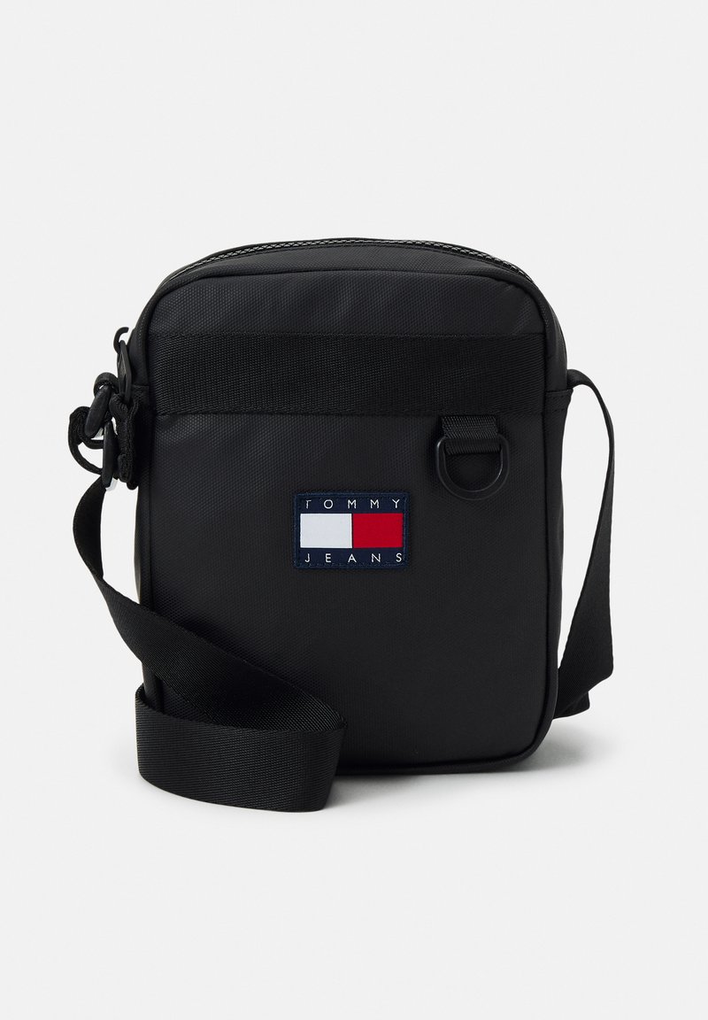 Bolso cruzado negro hecho de tela duradera, con cierre de cremallera y correa ajustable. Incluye un parche de logo rectangular en rojo, blanco y azul.