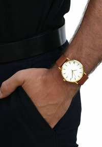 Bruin leren horloge met een ronde witte wijzerplaat, een gouden kast en minimalistische uurmarkeringen. Hand gepositioneerd in een broekzak.