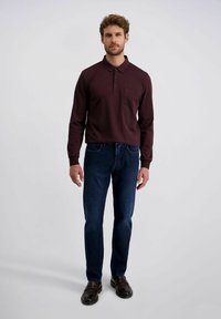 Man staat in een donker burgundy langmouw polo shirt, donkere spijkerbroek en bruine leren schoenen tegen een effen achtergrond.