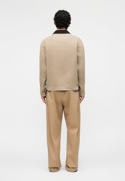 Άντρας με μαύρα σγουρά μαλλιά, φοράει μπεζ μακρυμάνικο πουκάμισο με μαύρο γιακά και χαλαρό μπεζ παντελόνι, στέκεται αντιμετωπίζοντας έναν λευκό τοίχο.