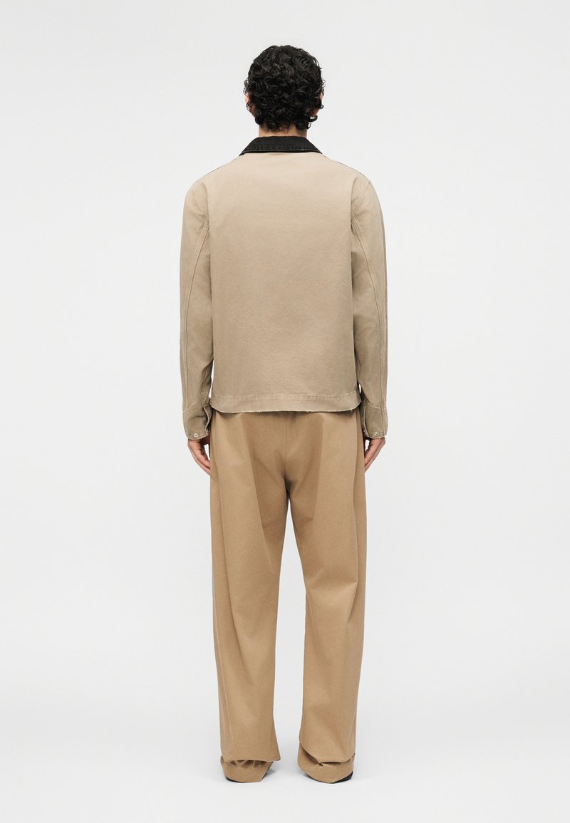 Man met zwart krullend haar, gekleed in een beige shirt met lange mouwen en zwarte kraag, en losse beige broek, staand met het gezicht naar een witte muur.