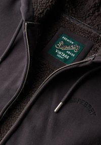 Sudadera con capucha negra con forro de borreguito, equipada con una etiqueta verde que dice "Superdry Vintage Sweat". Cierre de cremallera plateada y detalle del logo en relieve.