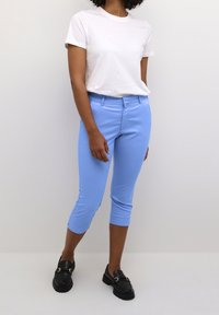 Pantaloni cropped blu chiaro in tessuto elasticizzato, con una parte frontale piatta e vestibilità slim, abbinati a una t-shirt bianca semplice e mocassini neri.