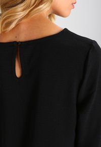 Czarny top z zaokrąglonym dekoltem i wycięciem w kształcie łezki z tyłu oraz subtelną fakturą. Długie rękawy i luźny krój.