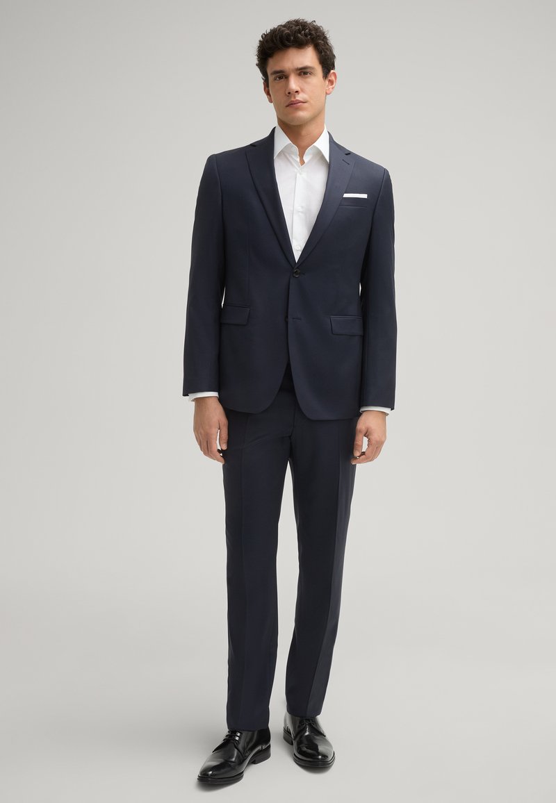 Abito blu con vestibilità su misura, chiusura a un bottone, rever a zadra e due tasche frontali. Abbinato con una camicia bianca e scarpe nere.