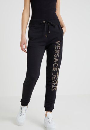 Kobieta nosząca czarne spodnie dresowe z napisem "Versace Jeans" z kryształków na lewej nogawce, z czarnym sznurkiem w talii, białe trampki i czarny top bez rękawów.