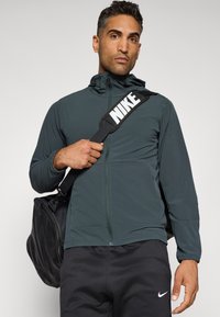 Mørkegrøn lynlåsjakke med høj krave, sammen med sorte bukser. Personen holder en sort taske med et fremtrædende hvidt Nike-logo på remmen.