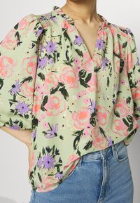 Blouse à fleurs en tissu vert clair présentant des motifs floraux roses et violets, un décolleté en V, et des accents froncés sur les manches.