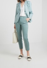 Blazer de teal claro con dos botones, combinado con pantalones ajustados a juego y una blusa blanca. Acabado con un bolso blanco y unos zapatos de slip-on con patrón.