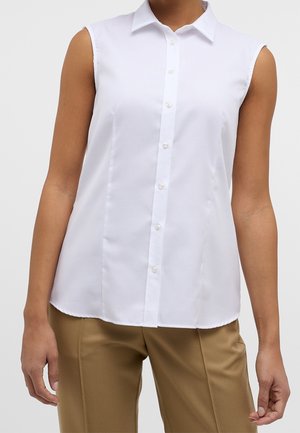 Mouwloze witte blouse met knopen, gedragen met beige broek, getoond van nek tot heupen tegen een effen achtergrond.