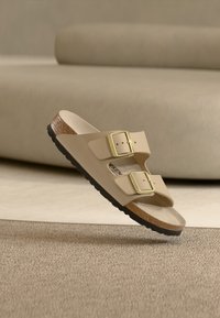 Μπεζ σανδάλι με δύο ρυθμιζόμενες χρυσές αγκράφες, φελλό πάτο και μαύρη σόλα, τοποθετημένο πάνω σε υφασμάτινο μπεζ χαλί και φόντο καναπέ.