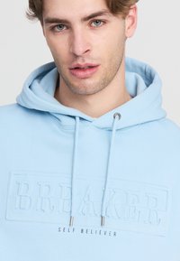 Felpa leggera blu chiaro realizzata in tessuto morbido, con un logo "BREAKER" testurizzato e in rilievo e un testo "SELF BELIEVER" in contrasto. Dettagli in metallo silver.