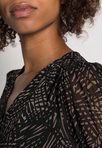 Schwarze Bluse mit einem geometrischen Muster in Beige, mit einem V-Ausschnitt und gerafften Schultern, aus einem leichten, durchsichtigen Stoff gefertigt.