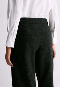 Pantaloni in lana nera a macchie con fascia in vita piatta e tasche posteriori. Camicia bianca con bottoni parzialmente visibile, con dettaglio ai polsini a sinistra.