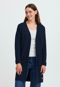 Long cardigan bleu marine en tissu doux, avec deux poches avant et un ourlet arrondi, porté sur un haut gris clair et un jean.