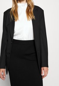 Černý blazer s klopami přes bílý top s vysokým krkem, zkombinovaný s černou perkovou tužkovou sukní. Elegantní, strukturovaný design, hladká textura.