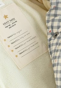 Krämfärgad fleece tyg med en vit etikett som har gyllene stjärnor och tryckt text, tillsammans med ett blått och vitt rutmönster.