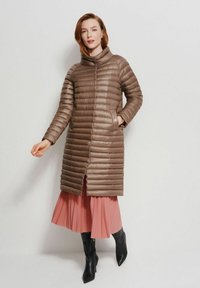 Brązowy pikowany płaszcz z wysokim kołnierzem, żebrową strukturą i poziomymi przeszyciami; zestawiony z różową plisowaną spódnicą i czarnymi kozaczkami sięgającymi do kolan.