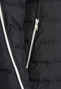 Czarna kurtka puchowa z diagonalnym wzorem pikowania, z kontrastowym białym zamkiem i uchwytem do zamka, co nadaje jej elegancki wygląd.