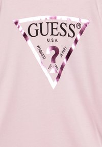 T-shirt di cotone rosa con un grande logo a triangolo in viola metallico e la scritta "GUESS U.S.A." e "WASHED JEANS" in nero.