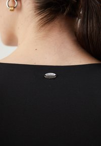 Czarna bluzka z okrągłym dekoltem, z metalową, owalną metką z napisem "WŁASNOŚĆ GUESS" przymocowaną z tyłu.