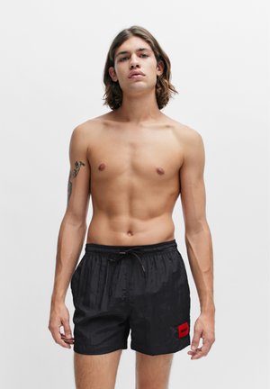Pantaloni da bagno neri realizzati in materiale leggero, dotati di una vita elasticizzata con cordoncino e una toppa con logo rosso sul lato sinistro.
