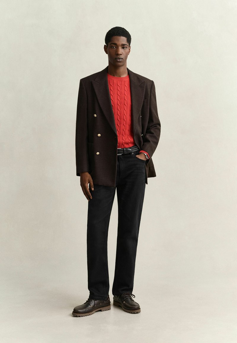 Young man wearing dark brown double-breasted blazer, red cable knit sweater, black pants, and brown lace-up shoes stands against plain background.