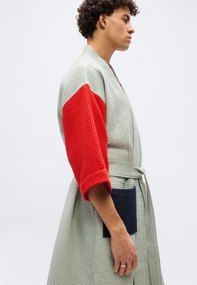 Νέος άνθρωπος που φοράει ρόμπα με υφή βάφλας, με γκρι κορμό, φωτεινό κόκκινο δεξί μανίκι, ναυτικό τσέπη και ζώνη στη μέση, στέκεται προφίλ μπροστά σε λευκό φόντο.