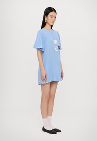 Helles blaues Baumwollkleid mit gewelltem Ärmel und Saum, verziert mit floralem Stickerei in Weiß und Schwarz. Kombiniert mit schwarzen Ballerinas und weißen Socken.