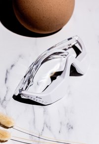 Białe sportowe gogle z przezroczystą soczewką, charakteryzujące się eleganckim designem i ochronną ramą. Zauważalny detal logo na boku; minimalistyczna estetyka.