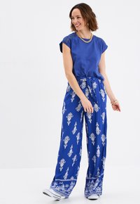 Top blu con maniche corte e dettaglio intrecciato sulle spalle. Pantaloni blu fluidi con motivo a fantasia e vita con nodo. Calzature casual.