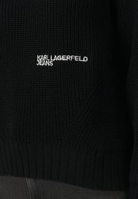Pulover negru tricotat, cu tiv și mansete cu dungi. Afișează "KARL LAGERFELD JEANS" brodat alb pe față.