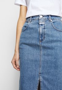 Person wearing a white t-shirt tucked into a high-waisted blue denim skirt with front pockets and a small front slit.