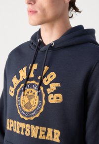 Navy blue sweatshirt with a hood, featuring yellow, distressed graphics that read "GANT 1949" and "SPORTSWEAR," and silver aglets on the drawstrings.