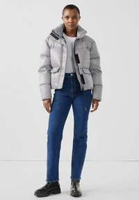 Chaqueta acolchada de cuadros blanco y negro con cuello alto, acentos negros contrastantes, bolsillos delanteros, combinada con vaqueros azules y botas negras.