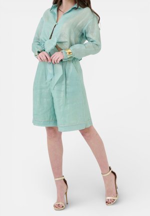 Donna che indossa una camicia a maniche lunghe verde chiaro con bottoni, annodata in vita, pantaloncini a vita alta abbinati, braccialetto d'oro e sandali beige con tacco alto.