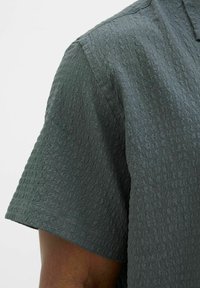 Košeľa s krátkym rukávom v tmavozelenej farbe, so štruktúrovaným vzorom a golierom. Materiál vyzerá ľahký s jemným, tkaným dizajnom.