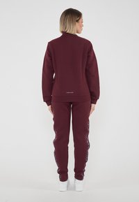 Bordowy sweatshirt i pasujące spodnie z miękkiego materiału, z wysokim kołnierzem, ściągaczami i wzorzystymi detalami po bokach. Białe trampki.