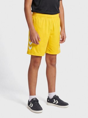 Barn iført lyse gule sportsshorts, sort skjorte, hvide strømper og sorte sneakers står på en ensartet lys baggrund.