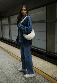 Übergroße Jeansjacke über einem grauen Hoodie, kombiniert mit weit geschnittenen Jeans und weißen Turnschuhen. Eine kleine beigefarbene Schultertasche wird getragen.