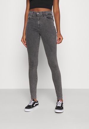 Femme portant un jean skinny gris taille haute, un top court noir et des baskets noir et blanc, debout devant un fond clair uni.