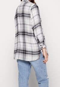 Camicia a quadri bianca e nera realizzata in tessuto morbido, con maniche lunghe, polsini con bottoni e orlo leggermente curvato. Vestibilità generalmente oversize.