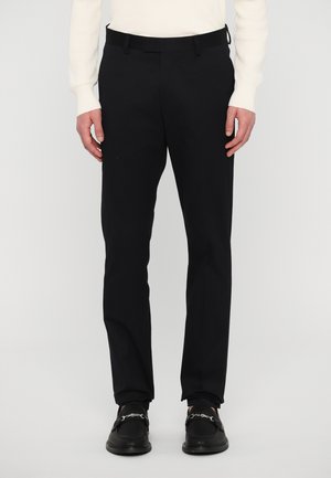 Calças pretas ajustadas com um corte slim, tecido liso e passantes para cinto. Combinadas com sapatos pretos tipo slip-on com detalhes em corrente decorativa.