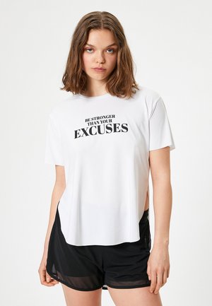 Hvid atletisk T-shirt med en afslappet pasform, der har sort tekst "BLIV STÆRKERE END DINE UDSKYLDNINGER." Sammen med sorte shorts, der har sideslids.