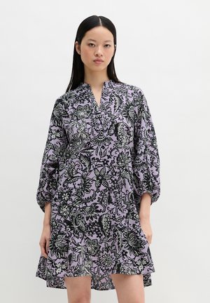 Femme portant une robe lilas ample avec des motifs floraux noirs et blancs, debout devant un fond blanc uni.
