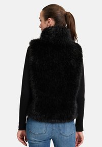Schwarze Kunstfellweste mit hohem Kragen, die eine weiche, strukturierte Oberfläche aufweist. Wird über einem Langarmshirt getragen und mit Jeans kombiniert.