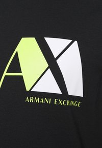 Svart t-shirt med en stor geometrisk logotyp i vitt och neongrönt, med texten "ARMANI EXCHANGE" nedanför i matchande neongrön stil.